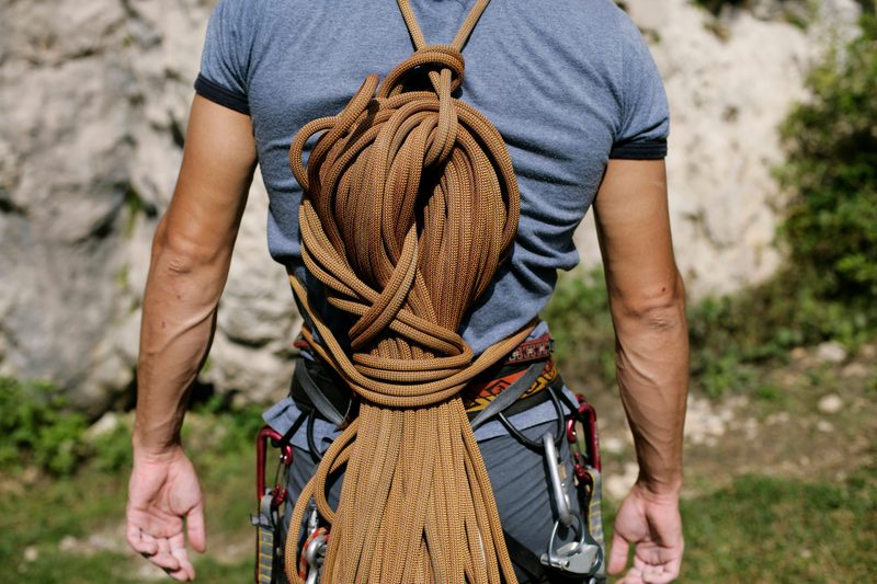 A climber seen from behind with a large coil of rope across his back and carabiners clipped to his harness, in front of a rock face.